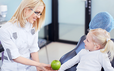 A young girl sitting in a dental chair with a dentist handing her an apple